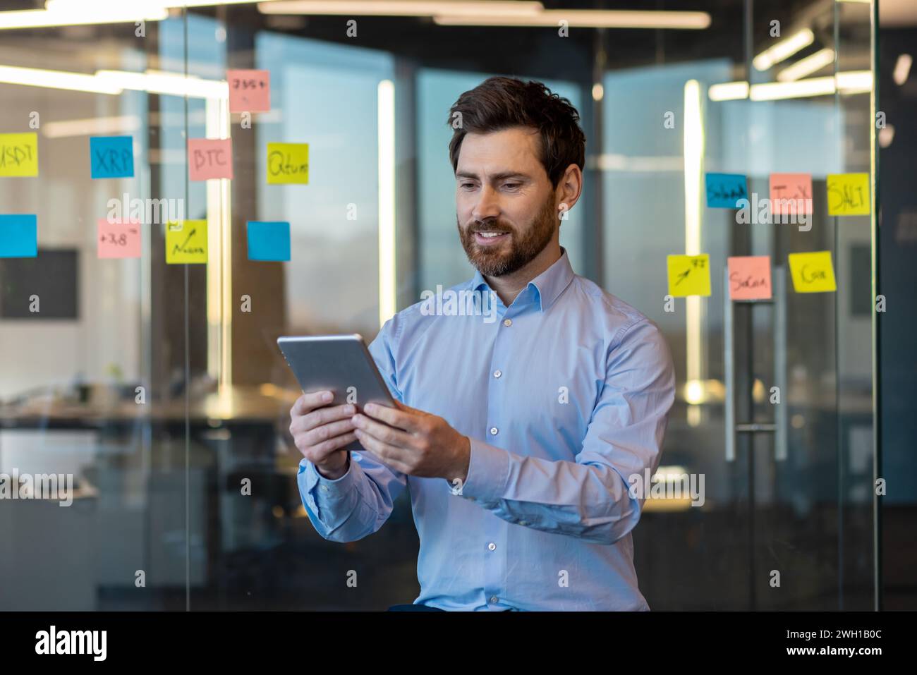 Mature businessman reviewing digital data on a tablet with sticky notes on glass wall in a well-lit contemporary office. Stock Photo