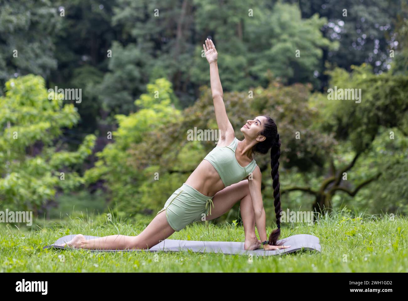Serene Indian woman in a green tracksuit practices yoga in a tranquil park setting, striking a pose on her mat among lush trees. Stock Photo