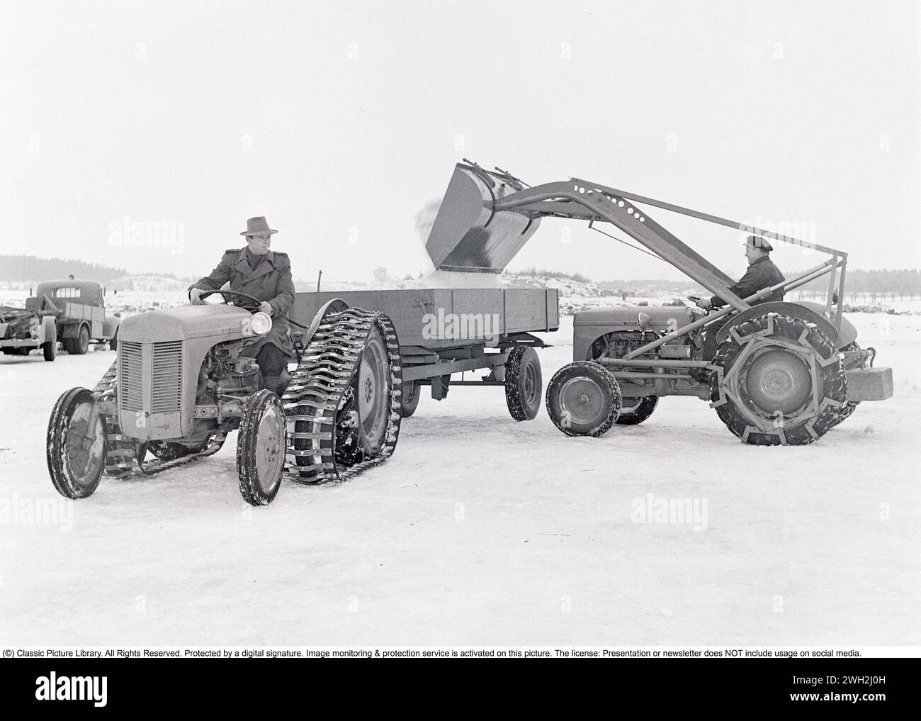 Ferguson tractor in the 1950s. A driver on the iconic Ferguson tractor featured with caterpillar bands and a one-axled wagon behind. Another Ferguson tractor with a loader, dumps snow onto it. The Ferguson tractors were the first modern agricultural tractors and its three poinkt linkage system was a major development.   1951. Kristoffersson ref BB46-2 Stock Photo
