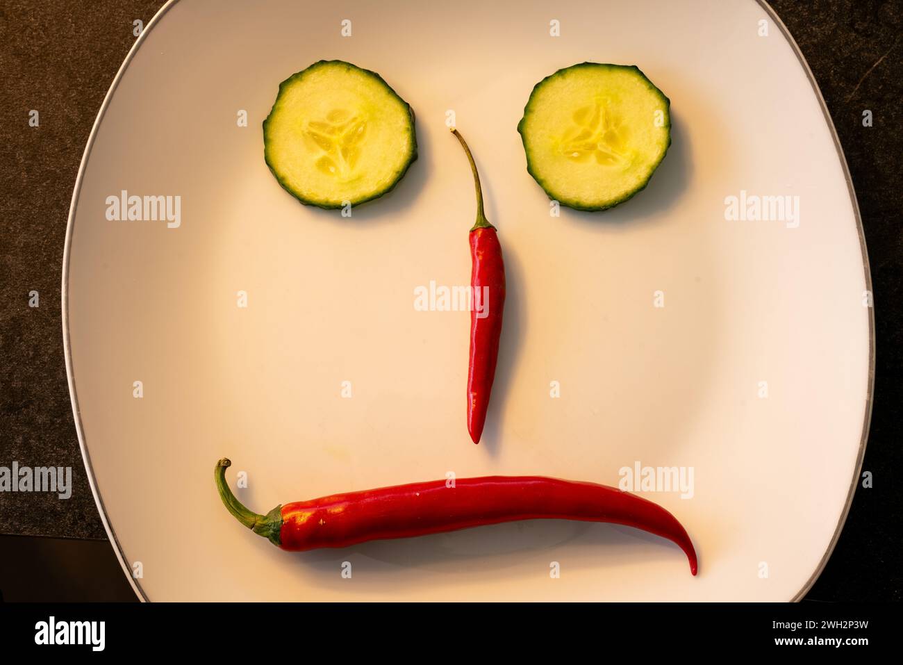 Frowning sad face arranged on a plate with red chili peppers and slices of snake cucumber. Stock Photo