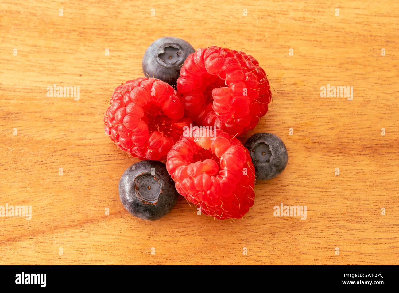 Close up of three raspberries and three blueberries arranged on a wooden snack board in a triangular shape. Stock Photo