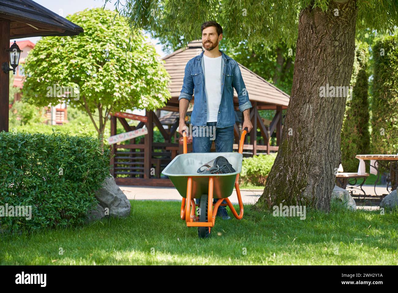Strong male gardener pushing wheelbarrow, while working in garden in sunny day. Front view of smiling man in denim shirt using cart for farming, while looking into distance. Concept of seasonal work. Stock Photo