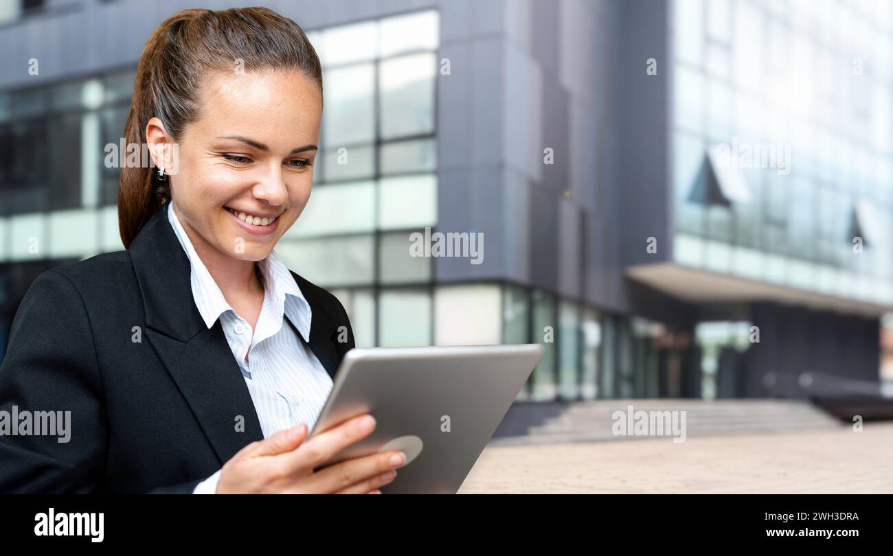 Young brunette businesswoman looking at digital tablet and smiling while standing on the street in front of business building. Stock Photo