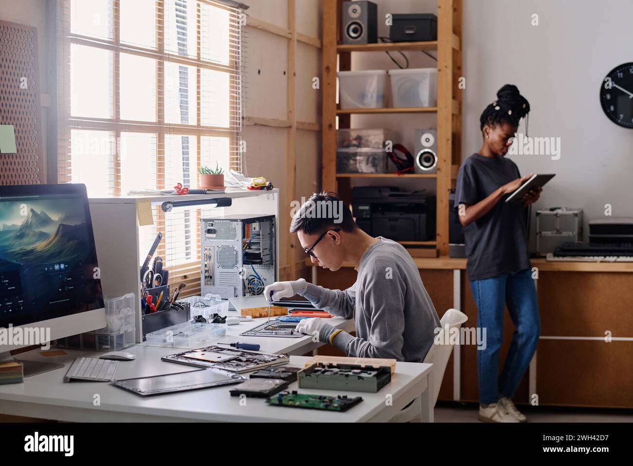Young repairman fixing broken phone sitting at desk in workshop, his black colleague standing nearby Stock Photo