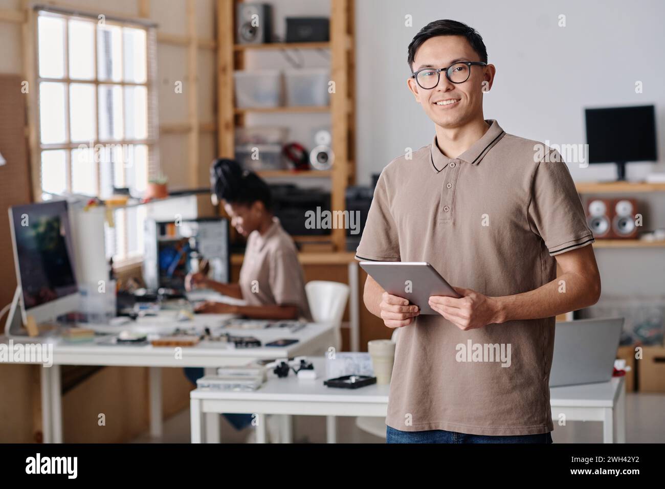 Male workshop owner confidently posing at workplace, black female technician working on background Stock Photo