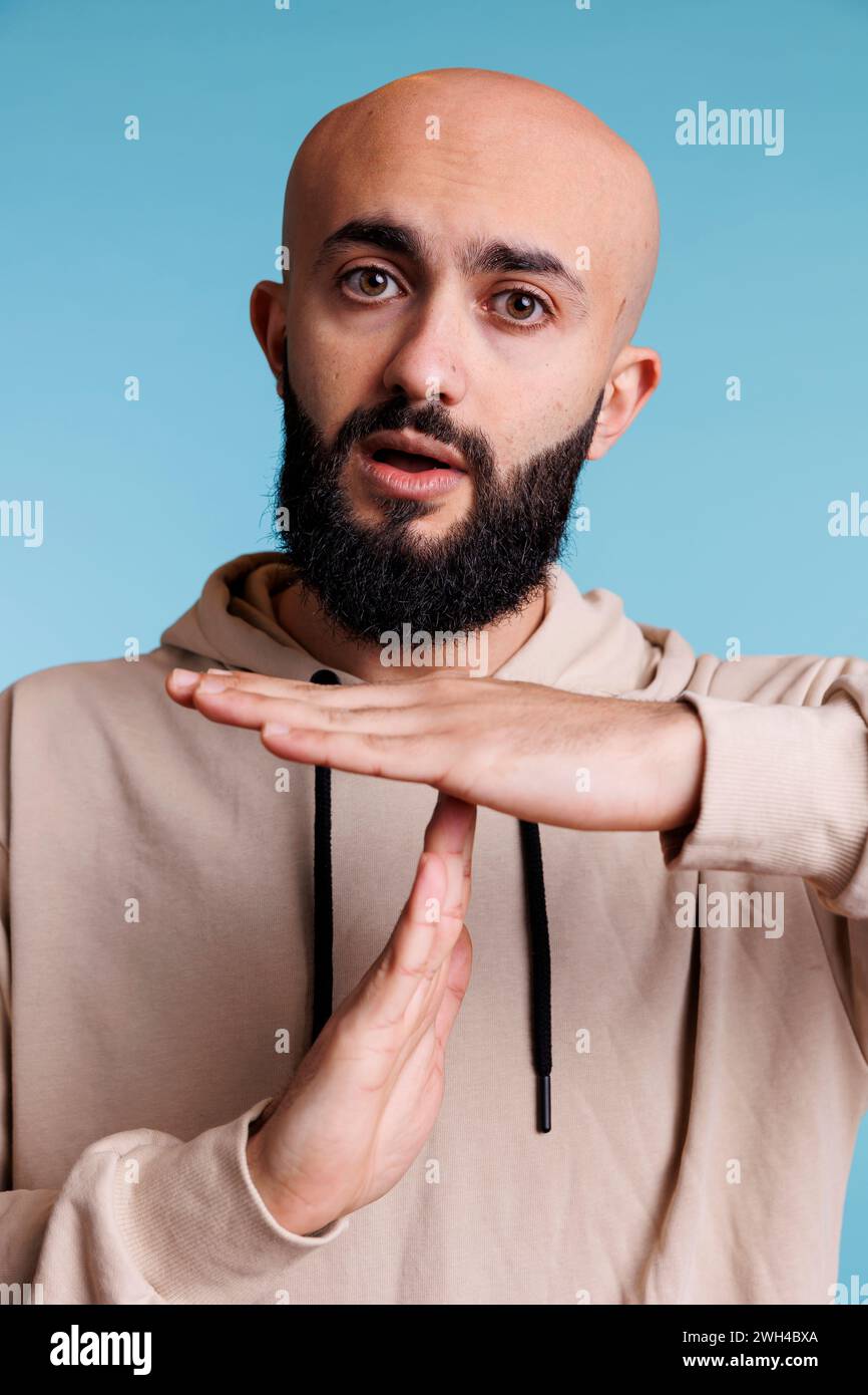 Arabian person taking time out with stop signal while showing confident facial expression portrait. Young bald bearded man making interruption gesture with hands while looking at camera Stock Photo