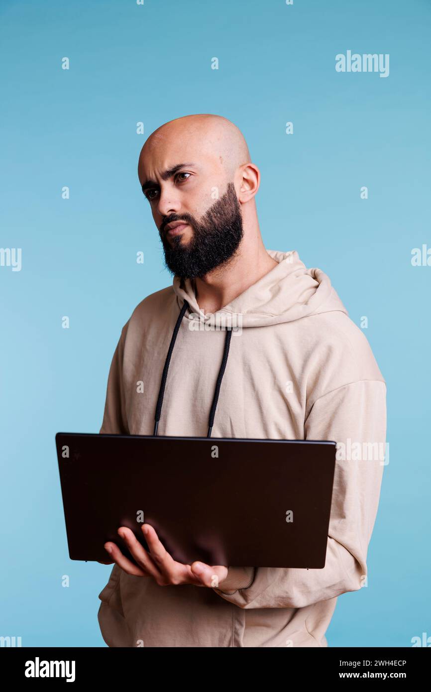 Puzzled arab man programming on laptop and looking away with confused expression. Confused person in doubt while developing software app and writing code on portable computer Stock Photo