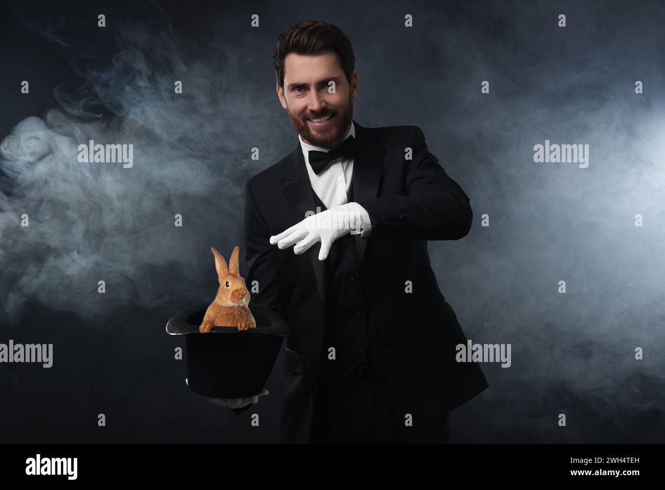 Magician showing trick with top hat and rabbit in smoke on dark background Stock Photo