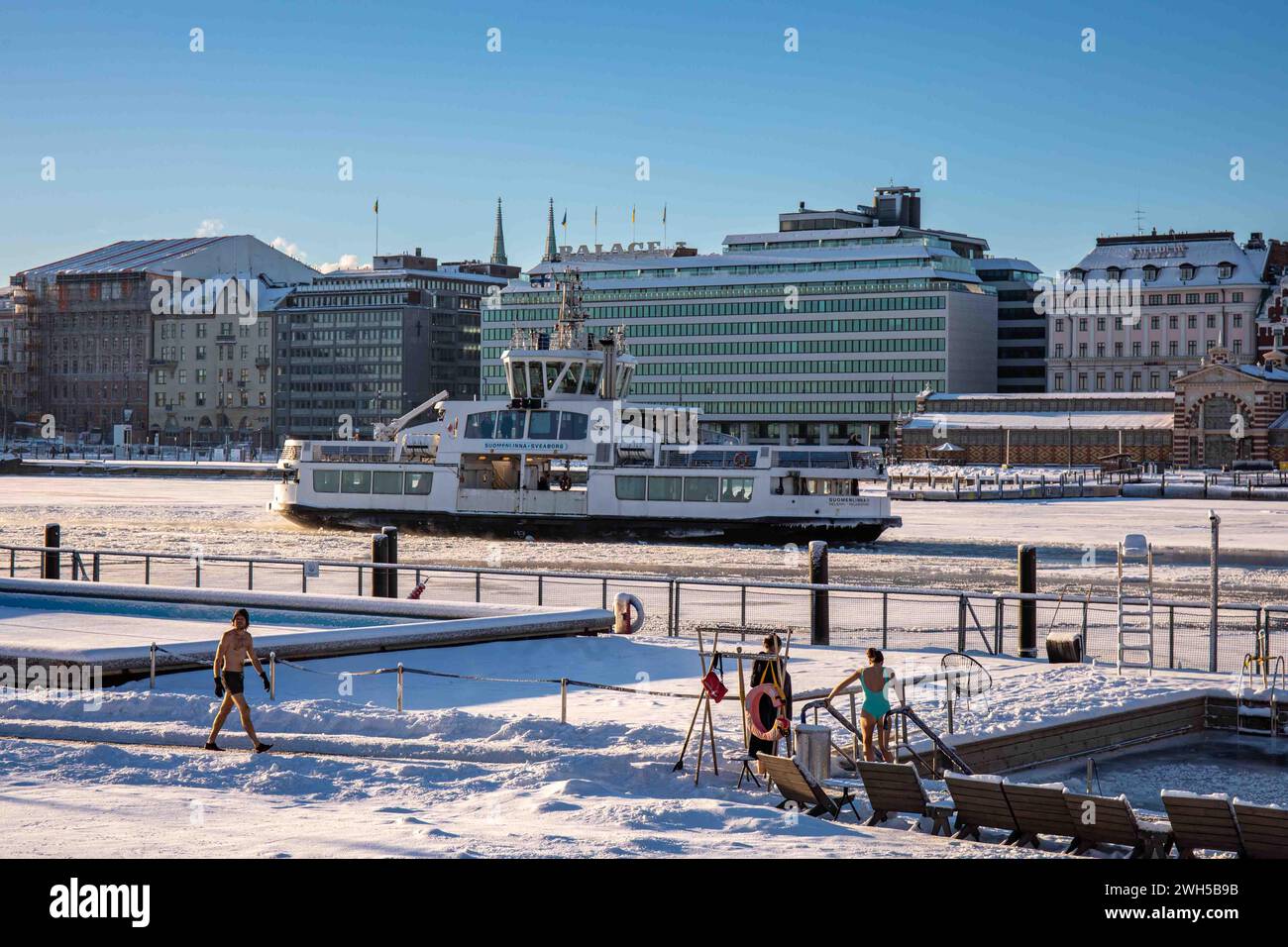 People on snow covered Allas Sea Pool floating deck with Suomenlinna II ferry in the background on a sunny winter day in Helsinki, Finland Stock Photo