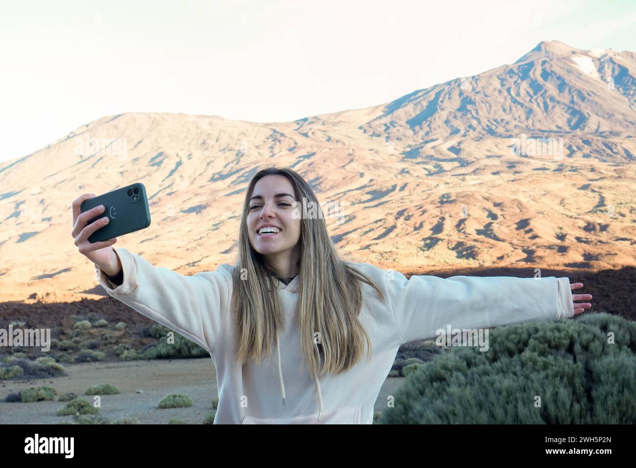 A joyful woman in a white hoodie is capturing a selfie with the majestic Teide mountain bathed in the warm glow of the evening sun. Stock Photo