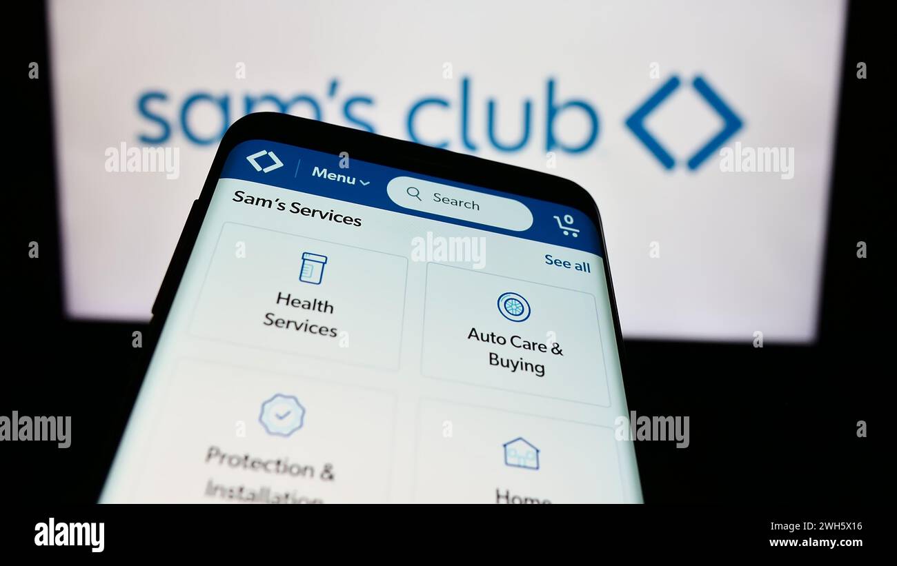 Mobile phone with website of US membership-only retail stores company Sam's Club in front of business logo. Focus on top-left of phone display. Stock Photo