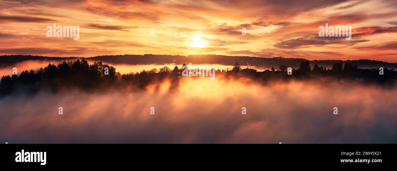 Dramatic sunrise over the fog and a range of tree silhouettes on a hill, a landscape panorama with dreamy sky, the rising sun and warm colors Stock Photo