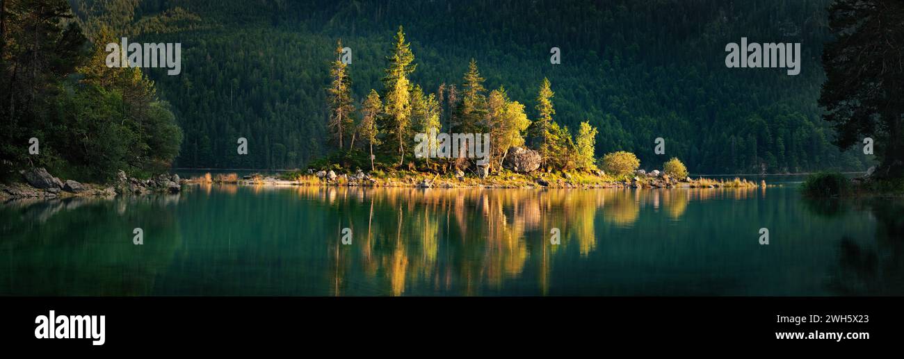 Gorgeous little island on a scenic lake lit by the sunset light and standing out from the dark mountain background, a wide panorama Stock Photo