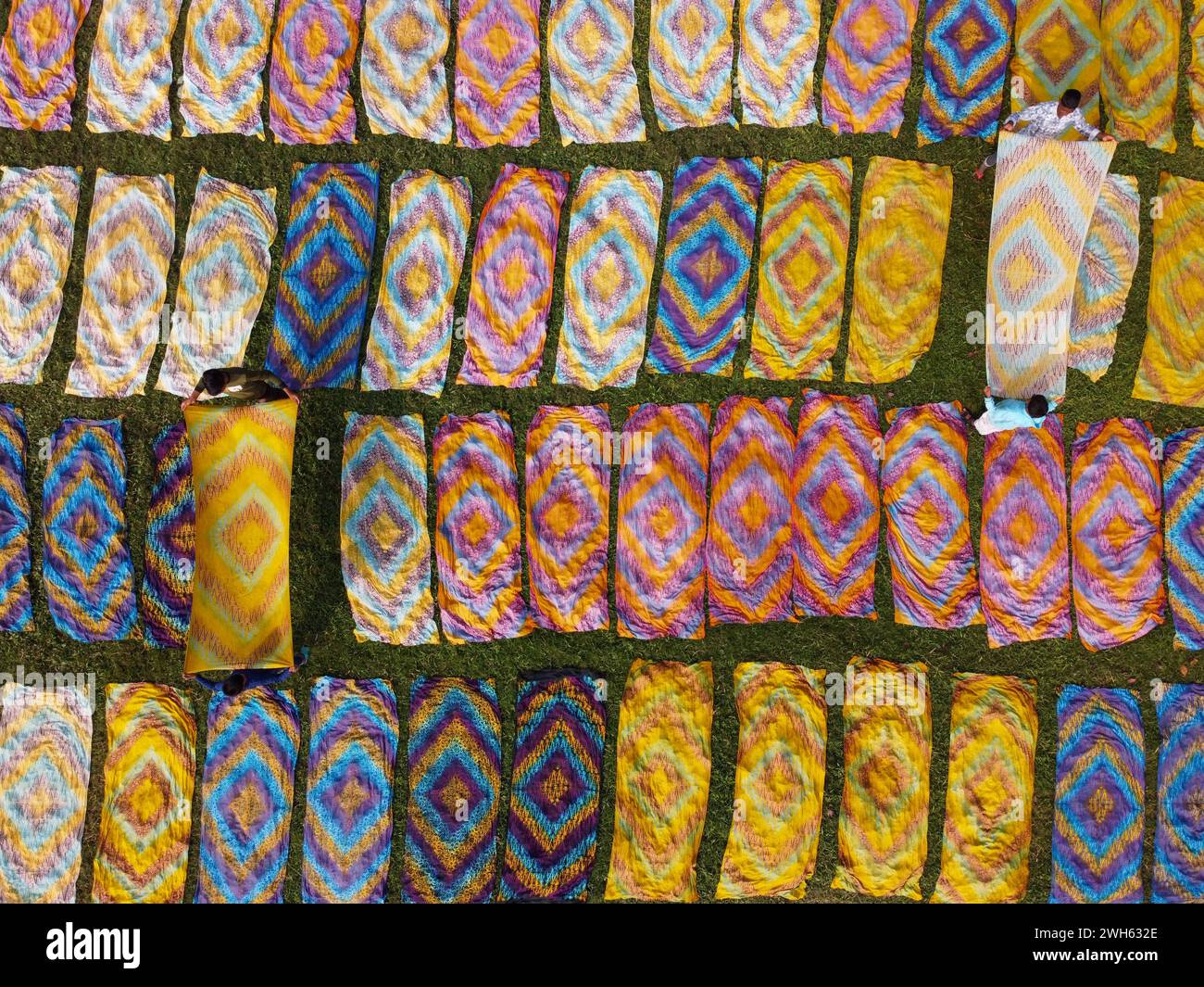 Narayanganj, Dhaka, Bangladesh. 8th Feb, 2024. Hundreds of pieces of dyed cloth are spread out across a field at Batik Village in Narayanganj, Bangladesh for drying - a process that usually takes 4 hours. The dyed fabrics, used in garment factories making T-shirts and women's dresses which sell for less than 3 Â£, are arranged on patches of grass to dry out completely. Once they are dried, they are made into clothes and sold worldwide. Beautifully embellished colorful cloths are created using the Indonesian technique called ''Batik''. Parts of the design are blocked out by applying hot wax Stock Photo