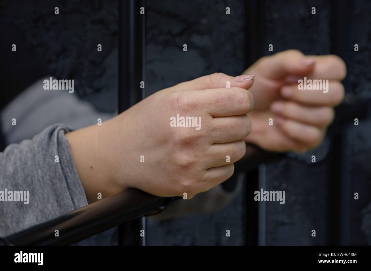 The woman's hands rest on the iron bars of the prison cell, restricting freedom. Stock Photo