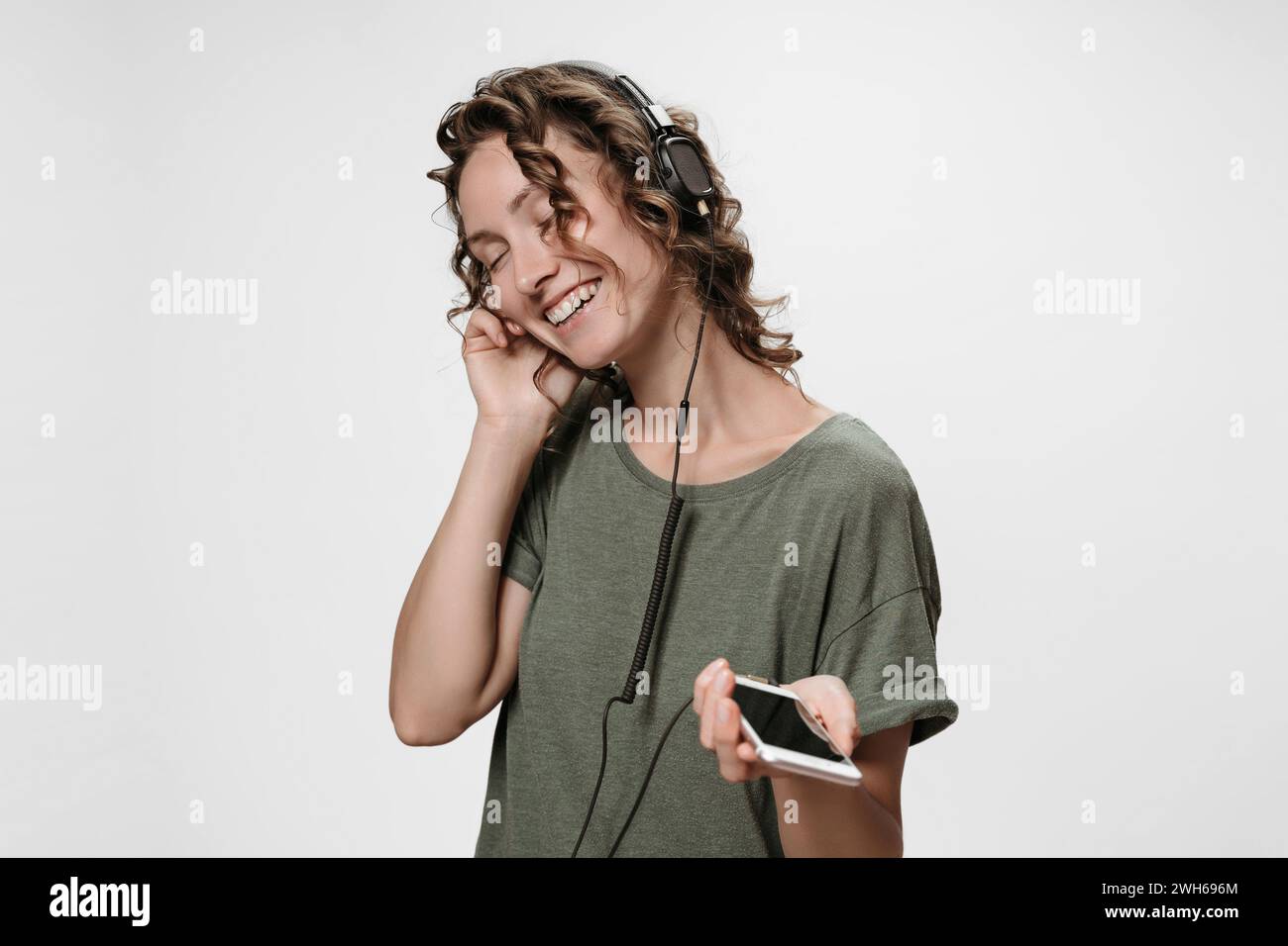 Carefree cheerful young curly woman listen favourite music with her stereo headphones, satisfied with quality of sound, enjoy favourite playlist, smiles, being in high spirit, isolated on white. Stock Photo