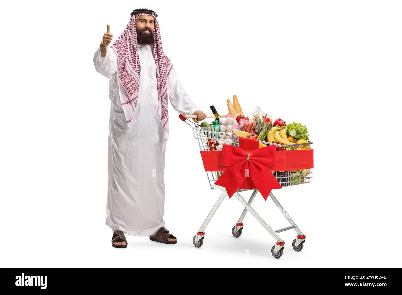 Arab man standing with a shopping cart full of food products and showing a thumb up sign isolated on white background Stock Photo