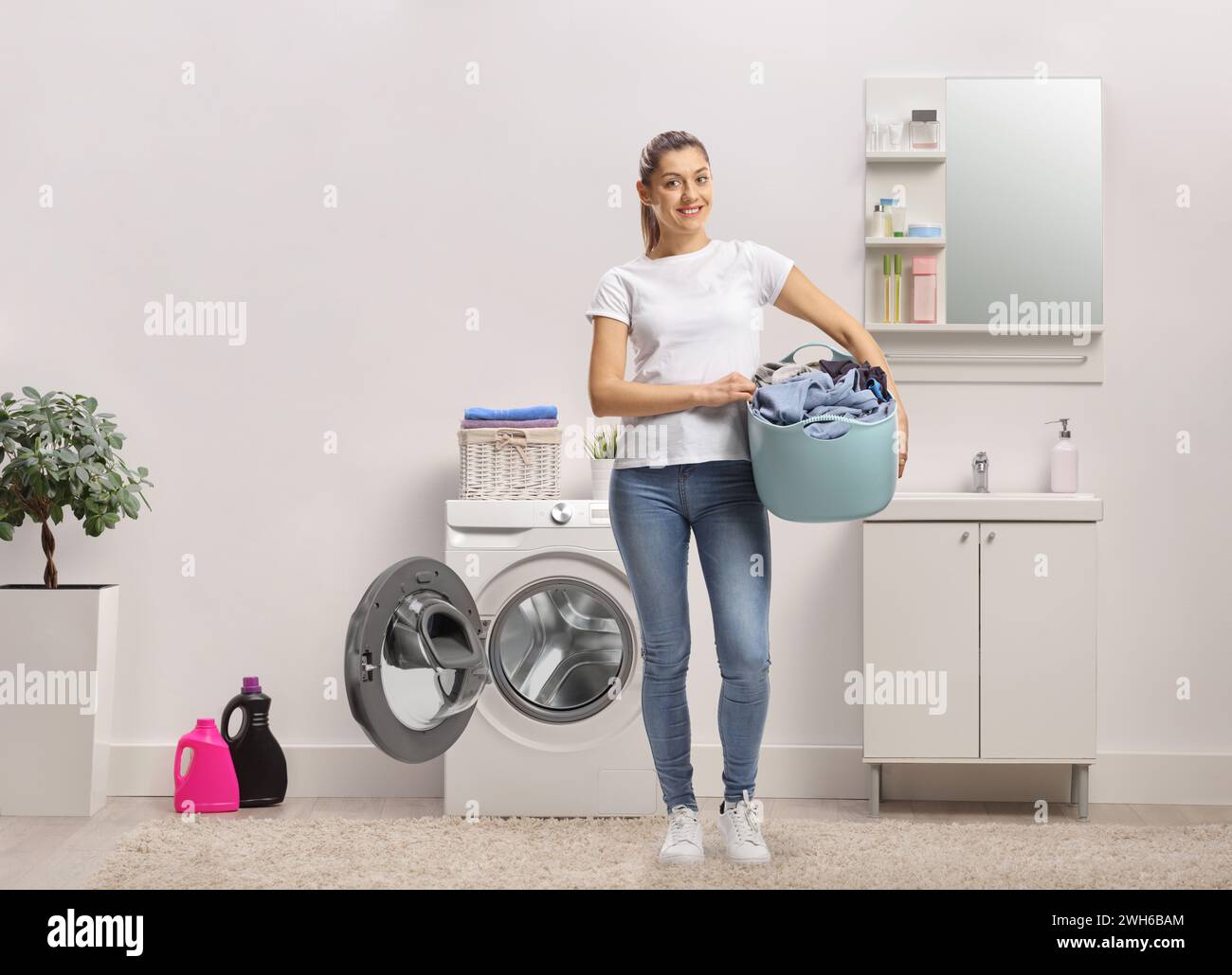 Woman in a bathroom holding a laundy basket next to a washing machine Stock Photo