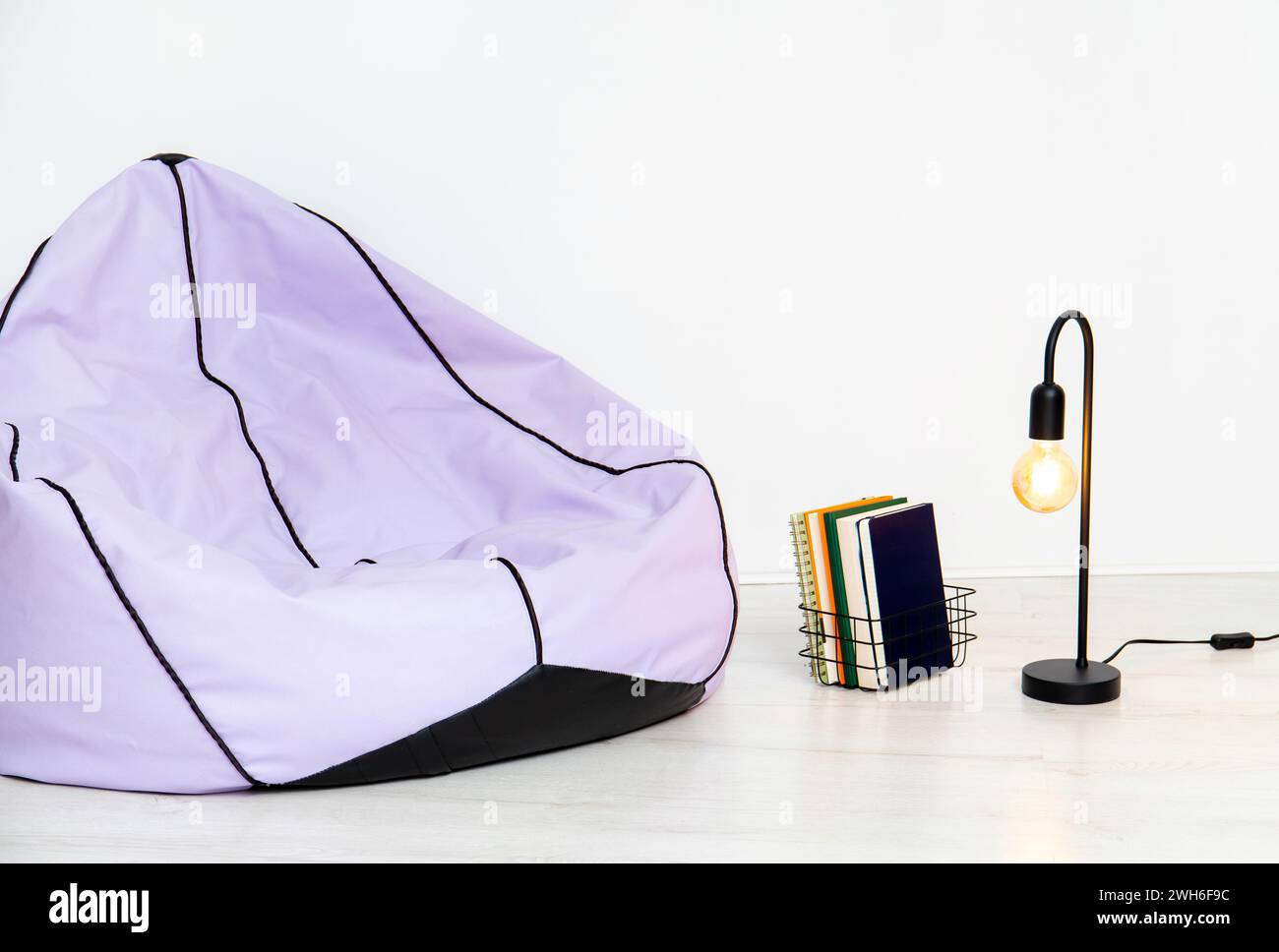 Minimalist home concept. Purple bean bag, black metal lamp and stack of books in metal wire basket, white walls and floor. Less is more. Stock Photo