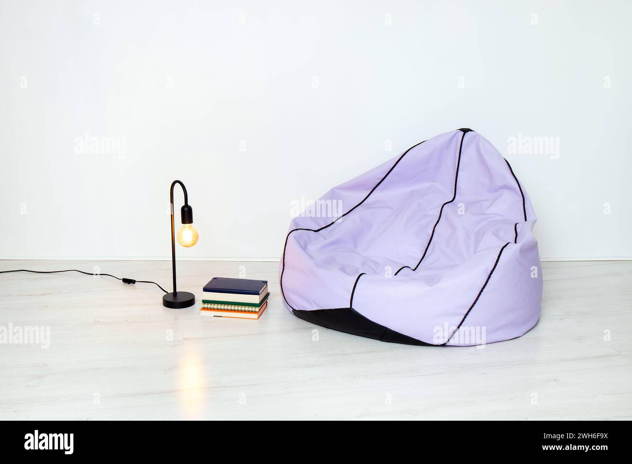 Minimalist home concept. Purple bean bag, black metal lamp and stack of books in metal wire basket, white walls and floor. Less is more. Stock Photo