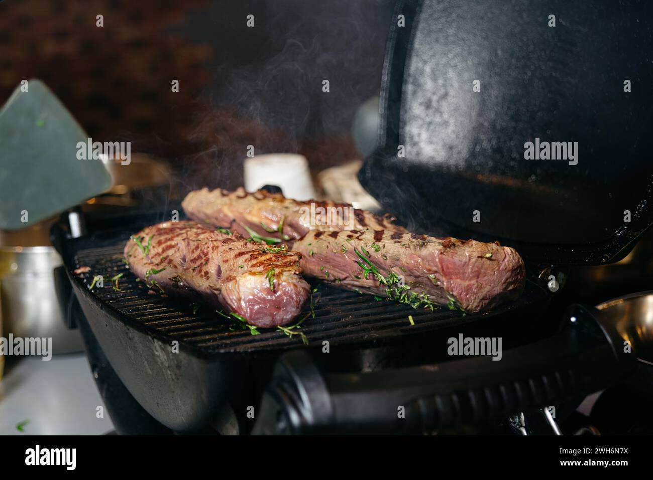 Juicy steaks seasoned with fresh herbs sizzling on a cast iron grill, steam rising in a warm, inviting kitchen setting. Stock Photo