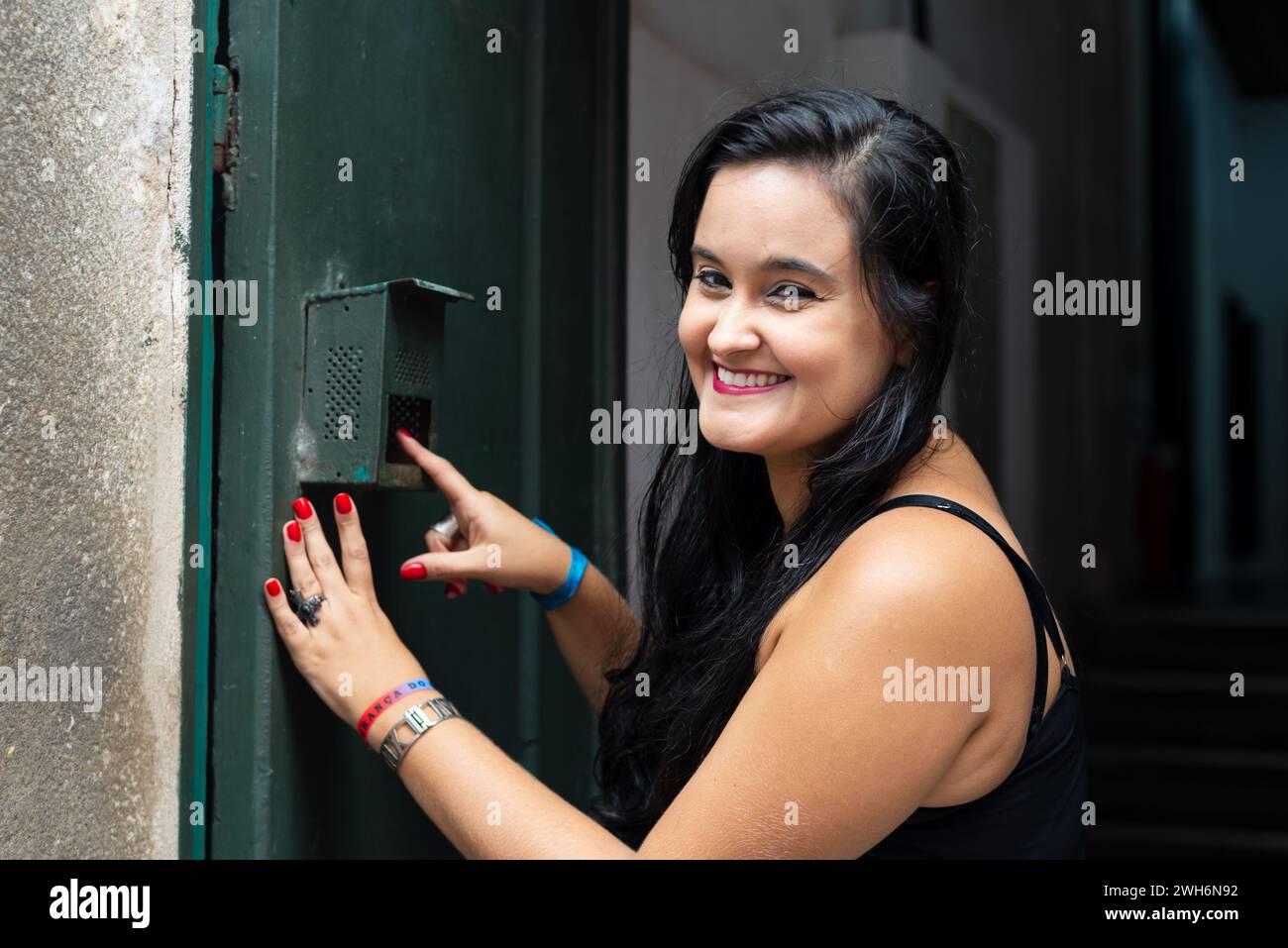 Portrait of beautiful young woman with black hair ringing the doorbell of a house and smiling. Person traveling. Stock Photo