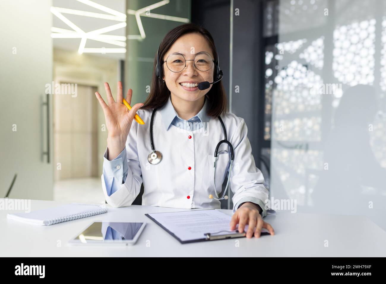 Smiling Asian female doctor in clinic office offers telehealth consultation, equipped with headset for remote medical assistance to patients. Stock Photo