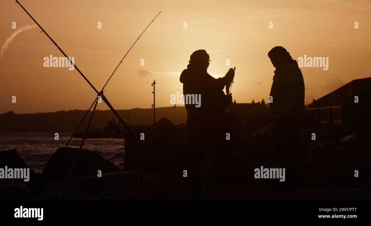 Silhouette Of Fishing Rods And Fishermen Measuring A Caught Sea Bass At