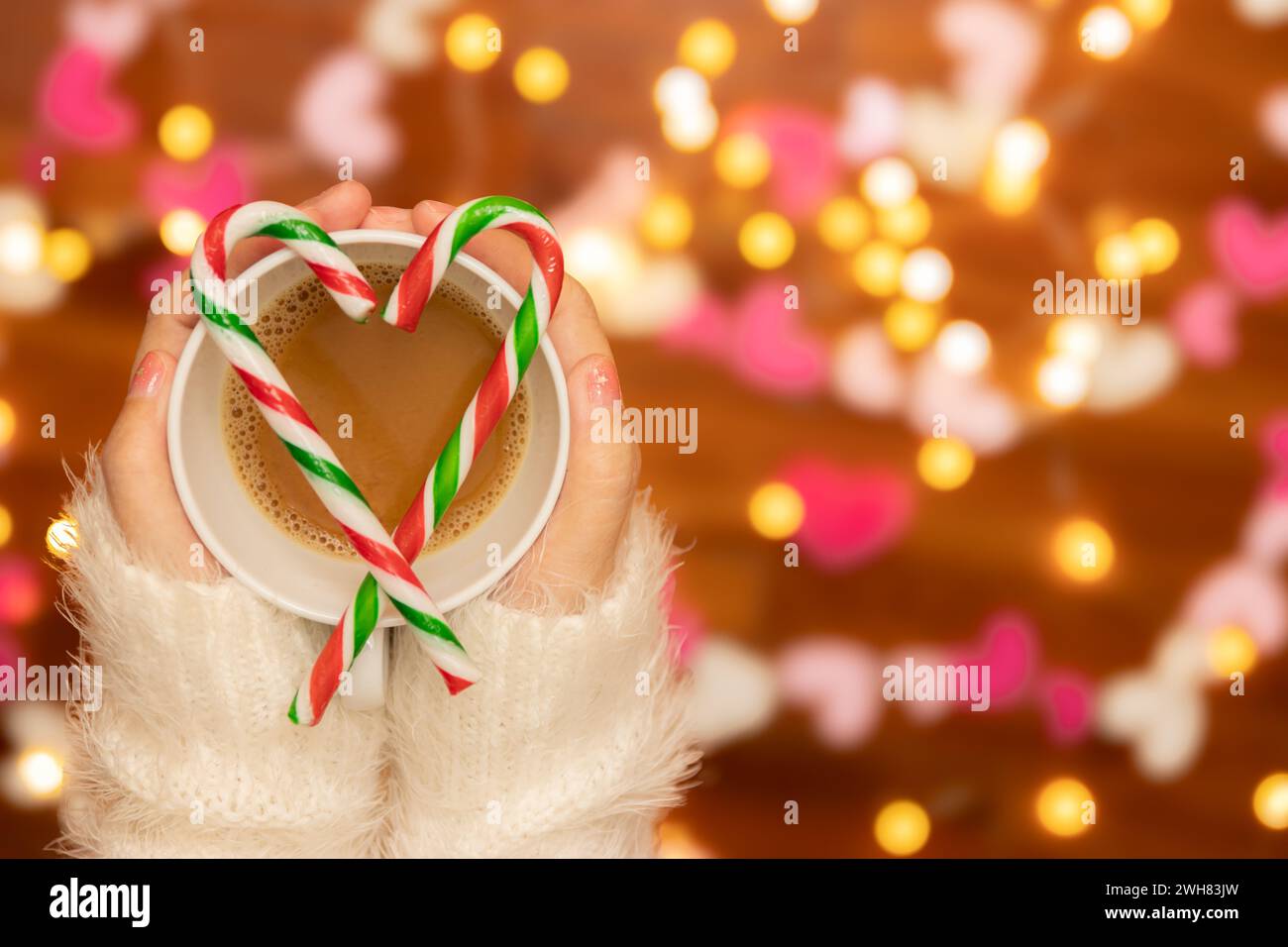 woman hand with sweater holding coffee cup with candy canes in the shape of heart on top, defocus bokeh light decorate for Valentine's Day celebration Stock Photo