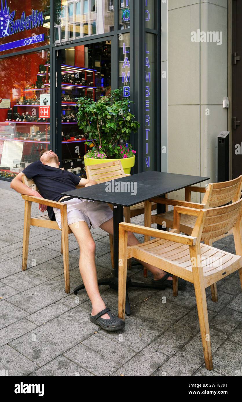 Young male traveler asleep at a cafe table in public view in downtown Cologne, Gemrany.  The weary traveler. Stock Photo