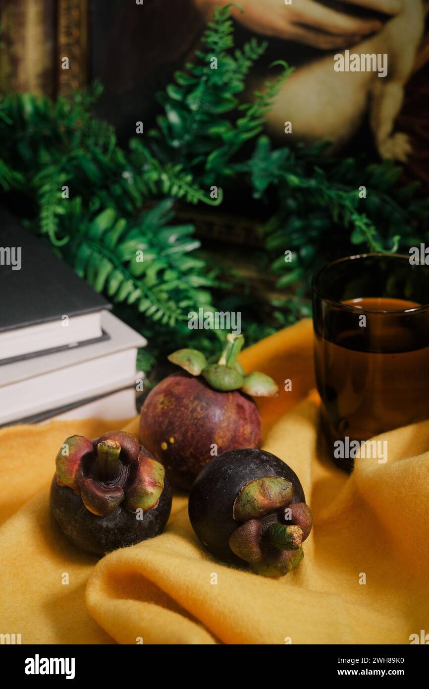 Lush still-life set up with three purple Mangosteens, books, vines, yellow felt cloth, a painting, glass of water. Close-up of sweet tropical fruit. Stock Photo