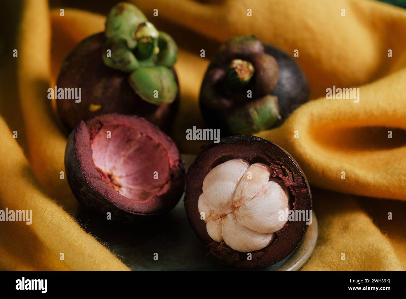 Lush still-life set up with yellow cloth, vines, a glass of water, and three purple Mangosteens, one cut open in half. Close-up of sweet tropical fruit. Stock Photo