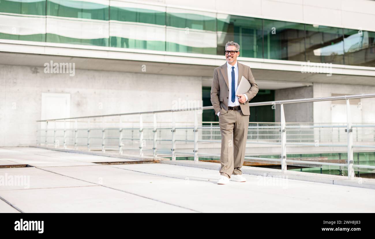 A full-length portrait of a mature grey-haired businessman smiling while walking confidently, laptop in hand, with a modern building in the background, suggesting a dynamic business lifestyle Stock Photo