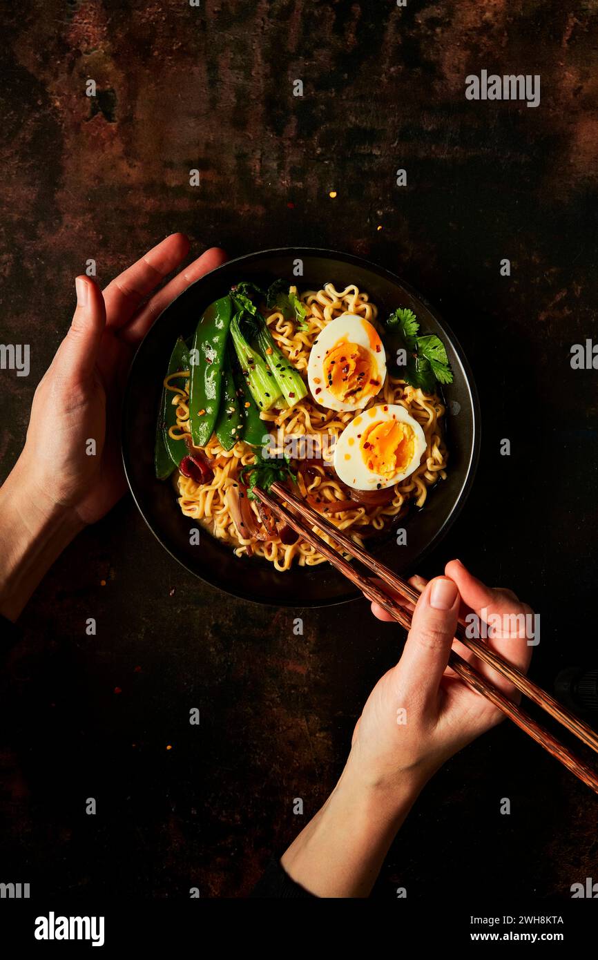 Hands holding chopsticks in Egg and Vegetable Noodles in black bowl with black napkin Stock Photo