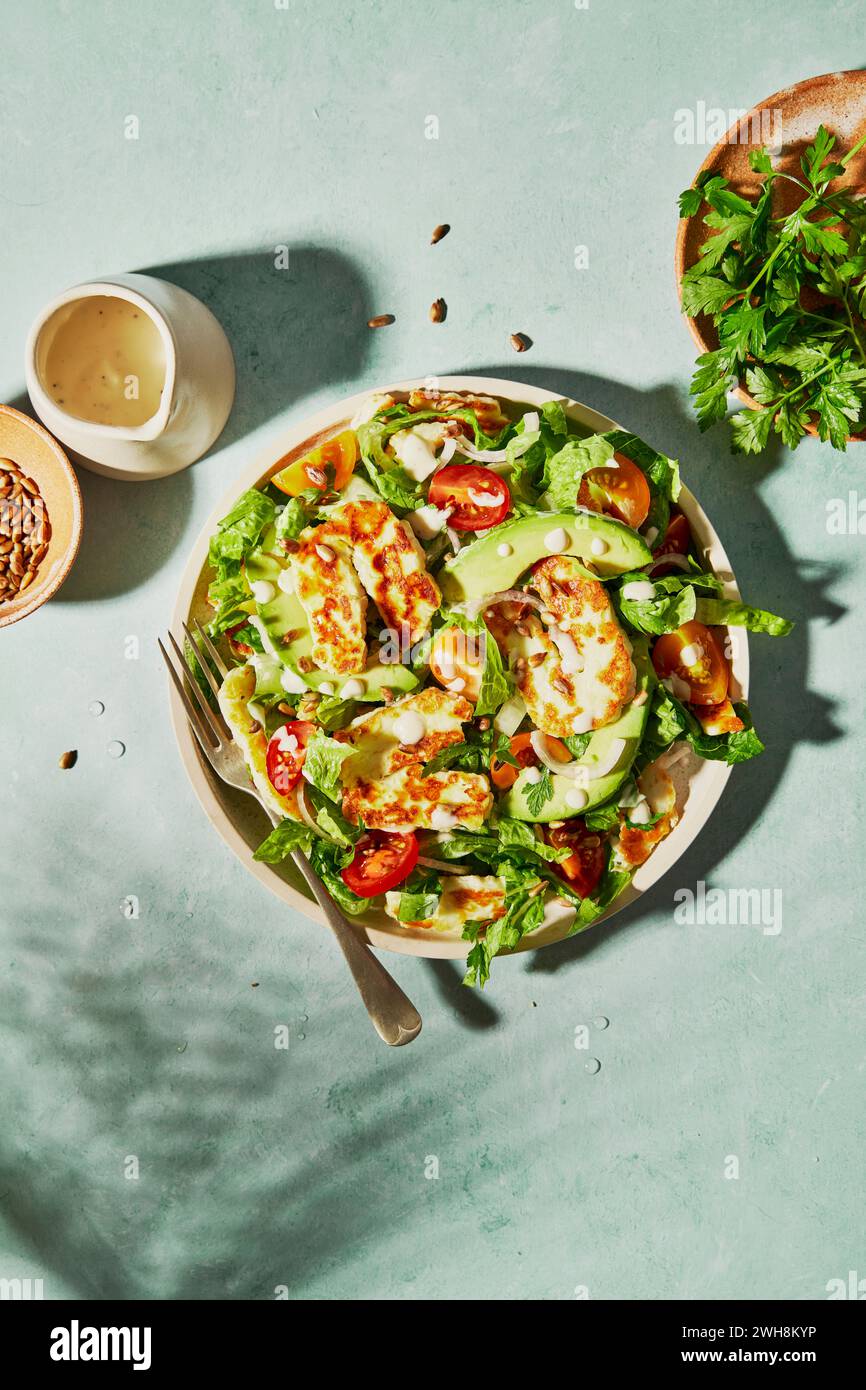 Halloumi Avocado Tomato Salad with sunflower seeds, yogurt dressing, herbs and water on a green background with shadow Stock Photo