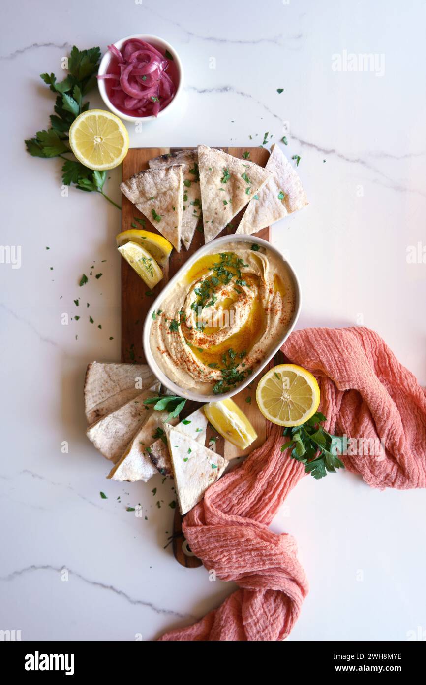 Chickpea hummus served with olive oil, pickled red onion, lemons, and flatbread on a white marble table background. Top view flatlay. Stock Photo