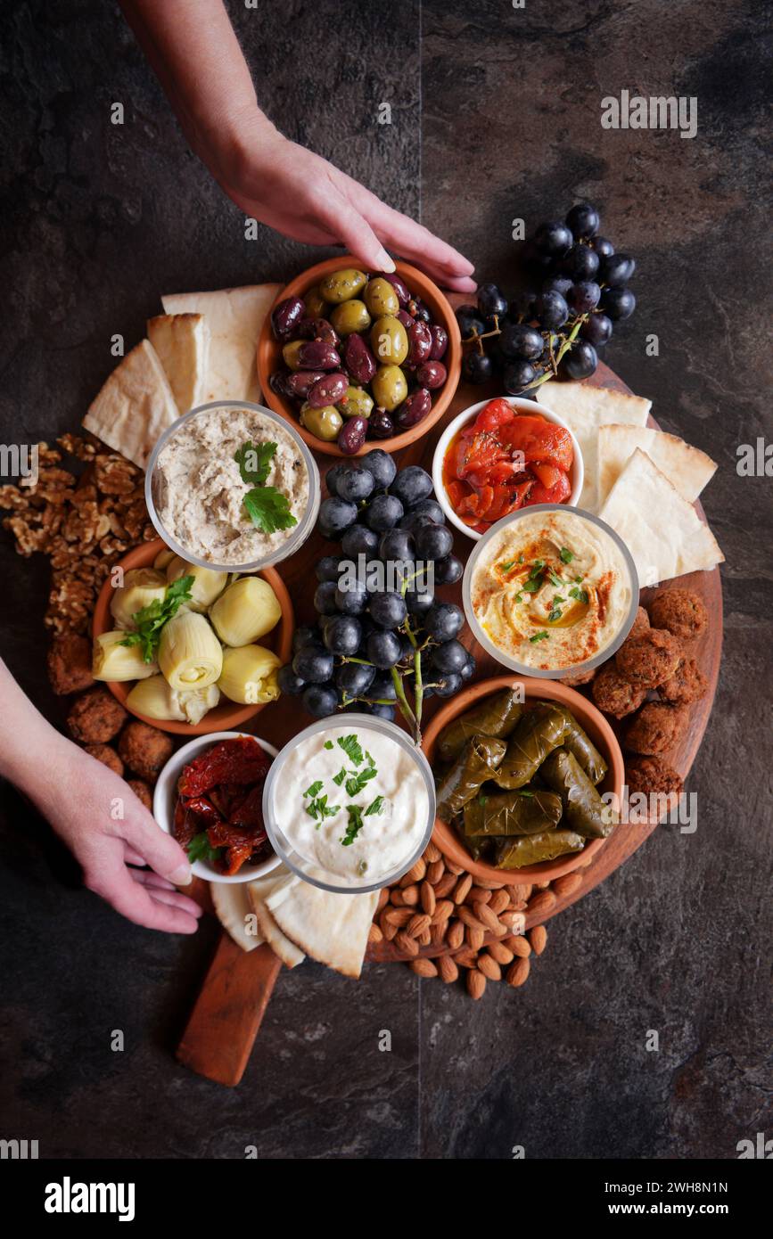 Female hands serving Mediterranean diet charcuterie board grazing platter on dark slate background. Stock Photo