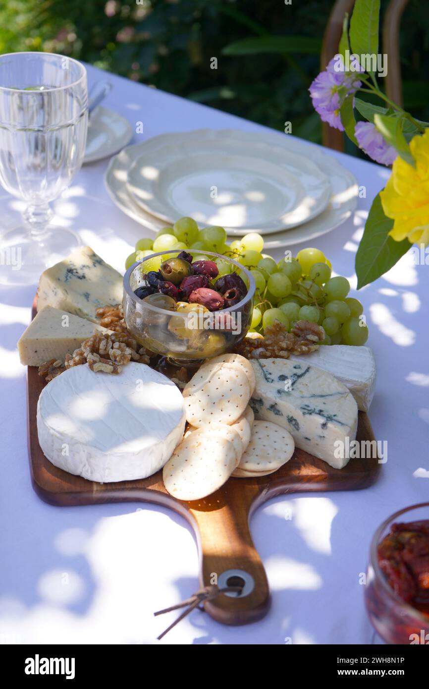 Al fresco summer outdoor dining table in dappled shade under a tall tree. Closeup on cheese and olives platter. Stock Photo