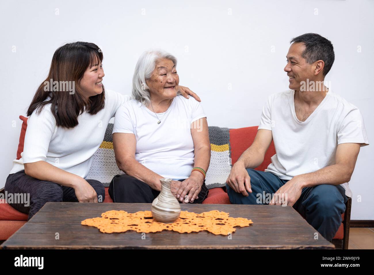 Adult children visit their mother of Japanese origin, chatting amicably sitting on sofa. Stock Photo