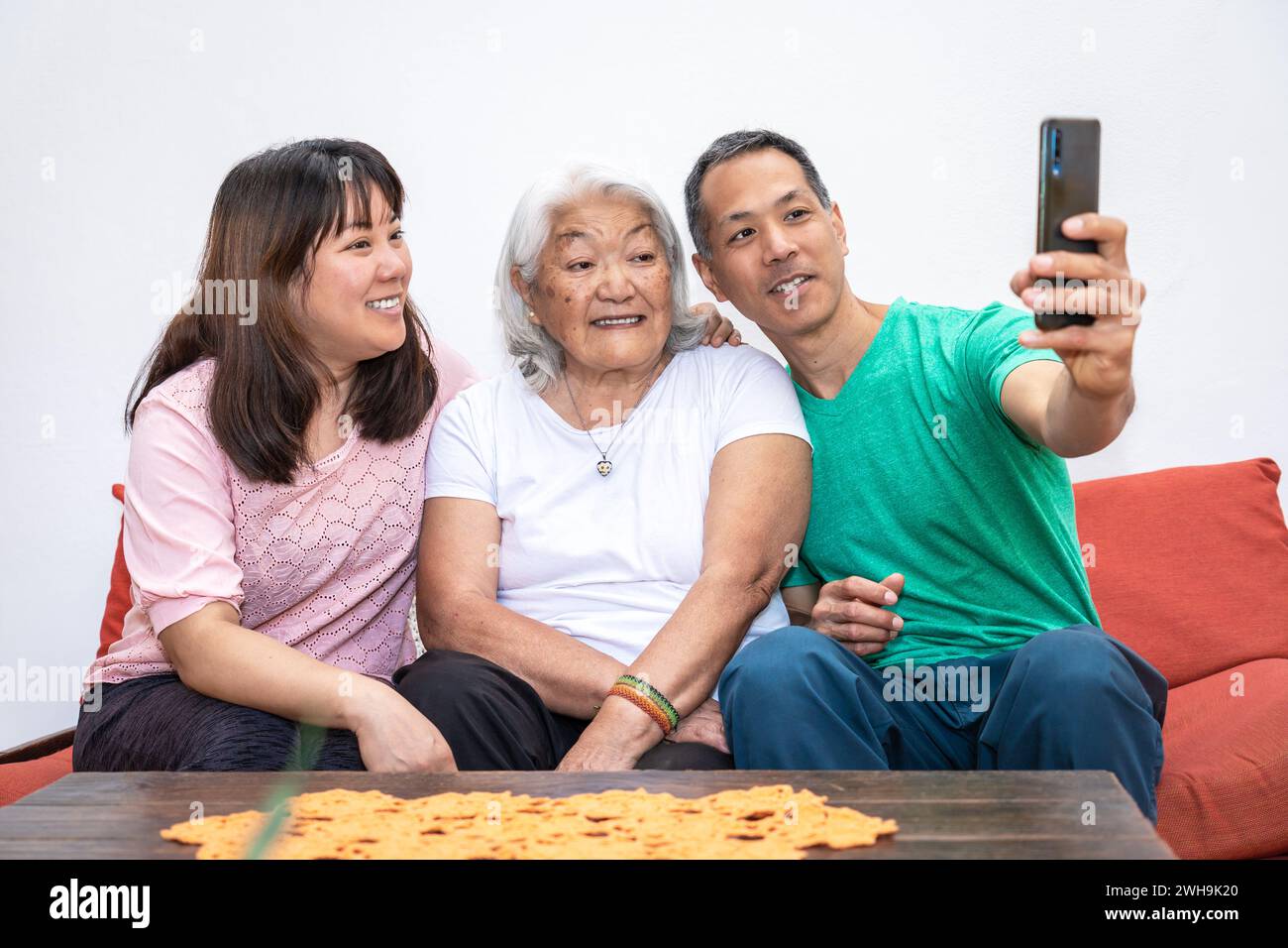 Adult children and elderly white haired mother, ethnic Japanese family, take a selfie sitting on a sofa smiling and sharing a family moment. Stock Photo