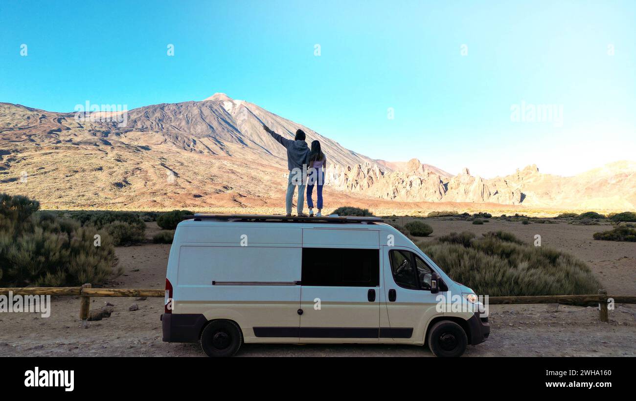 Two individuals standing on top of a van in the desert, as captured from a drone view in the Teide volcano mountain, Tenerife. Stock Photo
