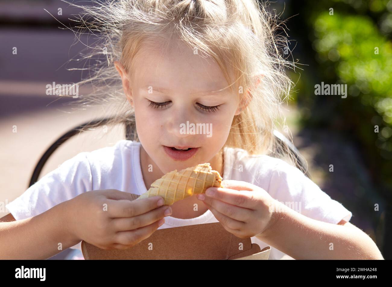 Little smiling girl have a breakfast in outdoor summer cafe. Cute caucasian girl eating tasty waffle tube Stock Photo
