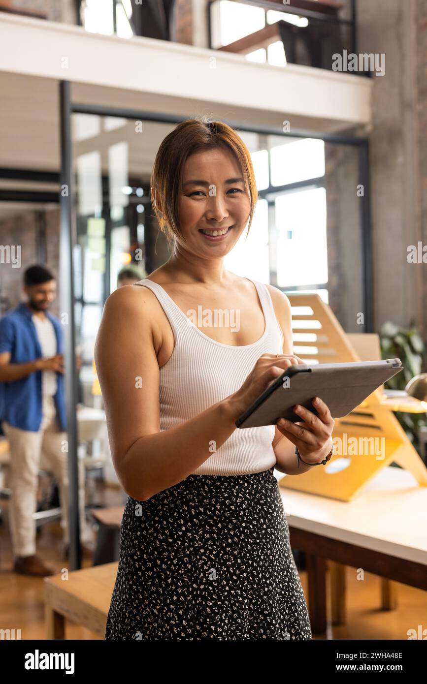 Asian woman in a casual business office setting, holding a tablet. She