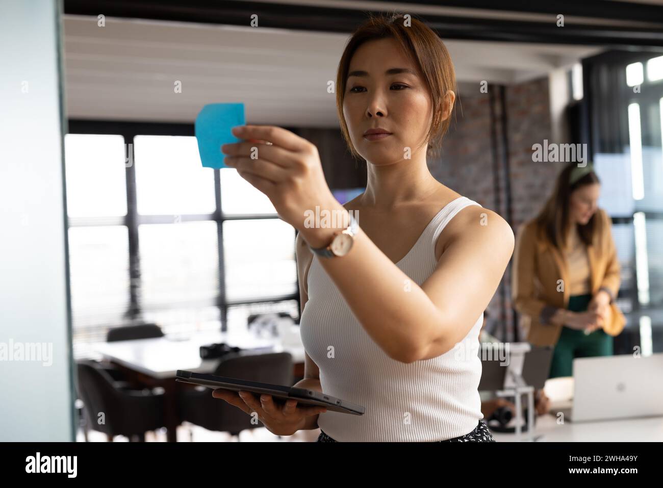 Asian woman reviews a sticky note in a modern, casual business office Stock Photo