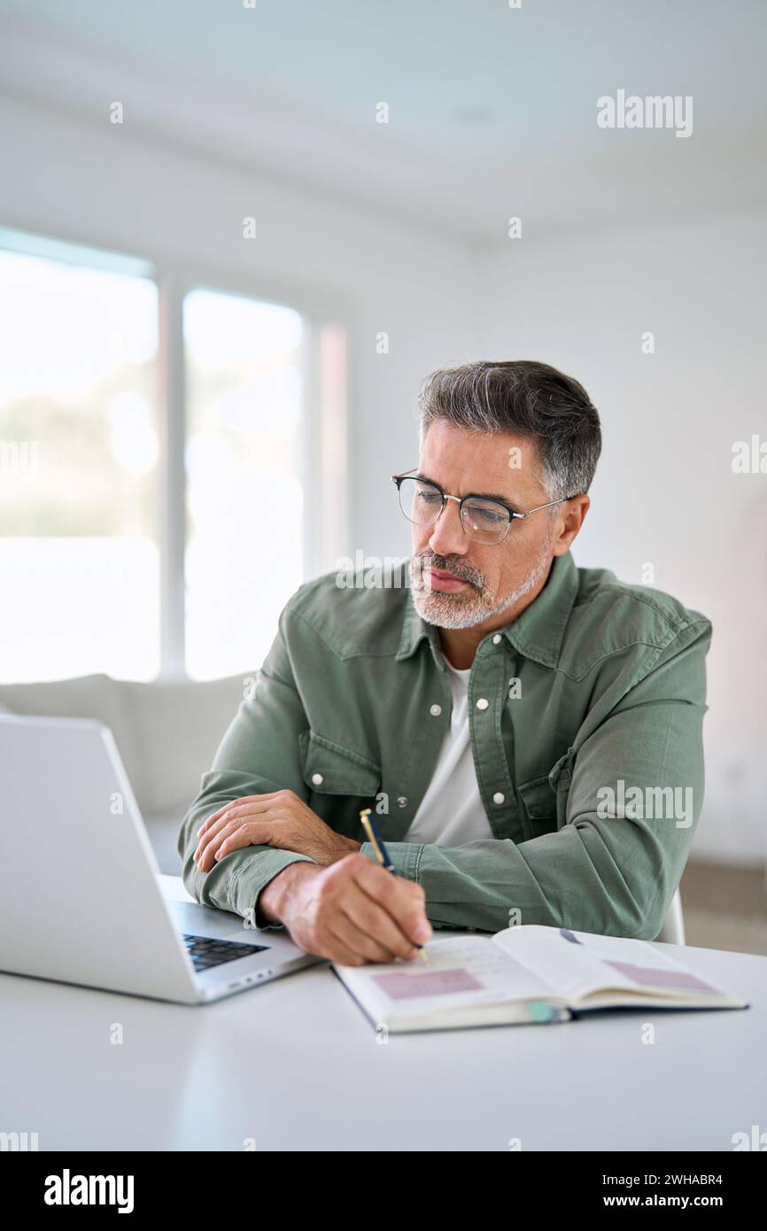 Serious older mature man using laptop computer writing notes at home table. Stock Photo