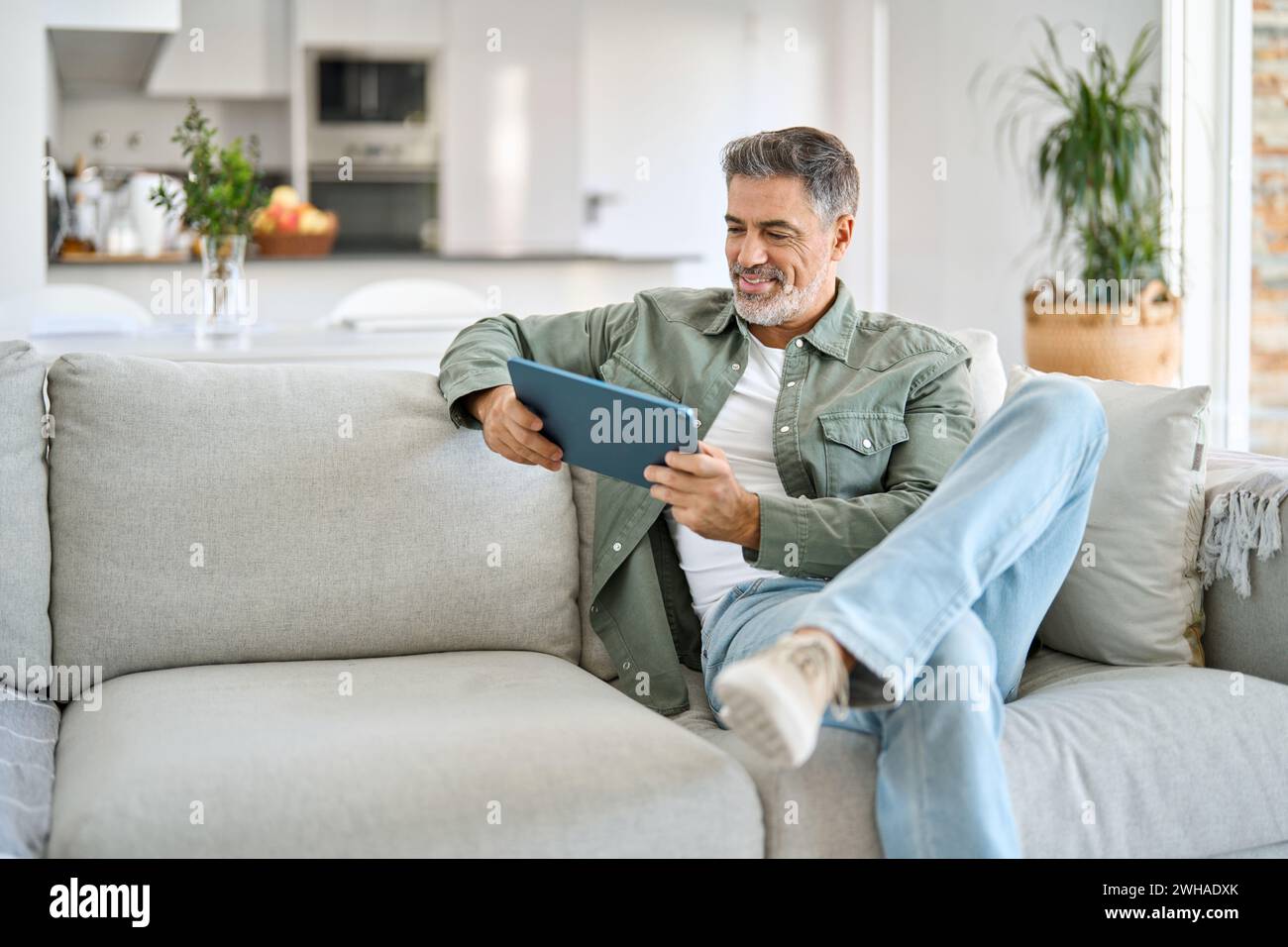 Happy middle aged man using digital tablet relaxing on couch at home. Stock Photo