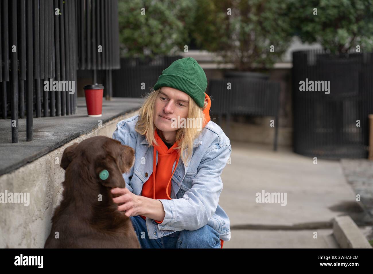 Kind warm-hearted stylish guy in trendy outfit sitting on haunches, caressing street dog with love Stock Photo