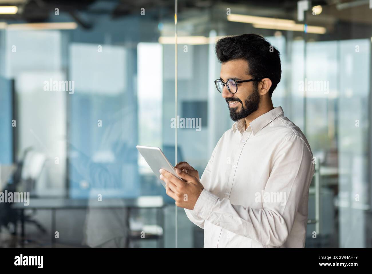 A man wearing a hat and beard stands by a glass wall, holding a tablet computer in his sleeve, displaying content using his thumb gesture. Stock Photo