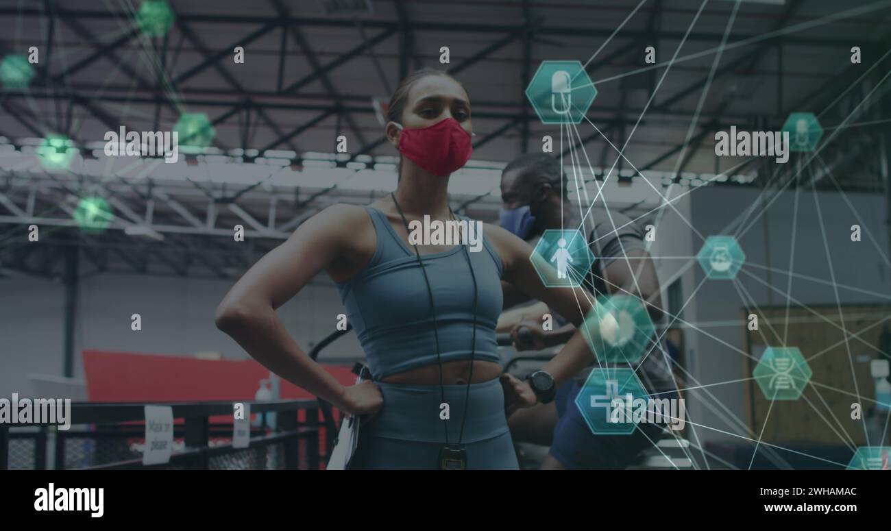 Globe of medical icons spinning against female fitness trainer wearing face mask standing in the gym Stock Photo