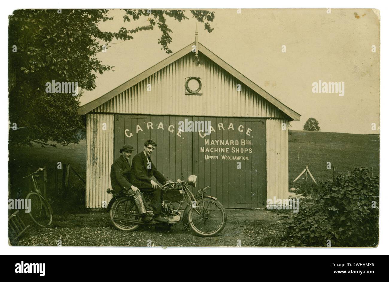 Original early 1900's postcard of two young lads, friends, sitting together on a motorbike outside Maynard Brothers Garage, Machine Shops, Upper Woolhampton, Berkshire, U.K.  The garage is constructed of corrugated tin / iron. A bicycle is propped up outside. One of the lads wears improvised goggles, the pillion passenger wears paint- splattered workman's trousers. Circa 1920's Stock Photo